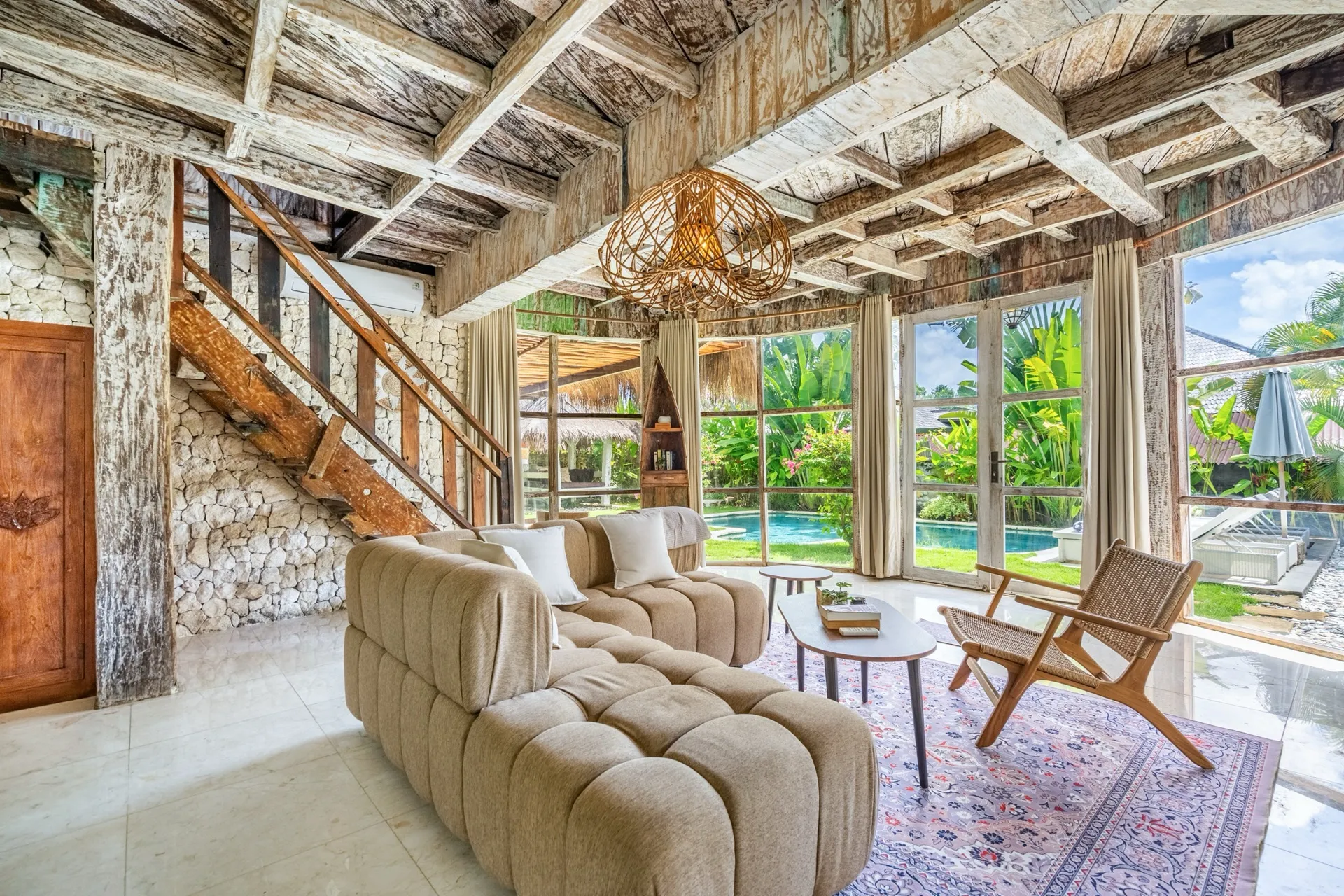 The living room at Villa Idaman with whitewashed reclaimed-timber beams, a hand-cut stone wall and glass doors looking out to the pool