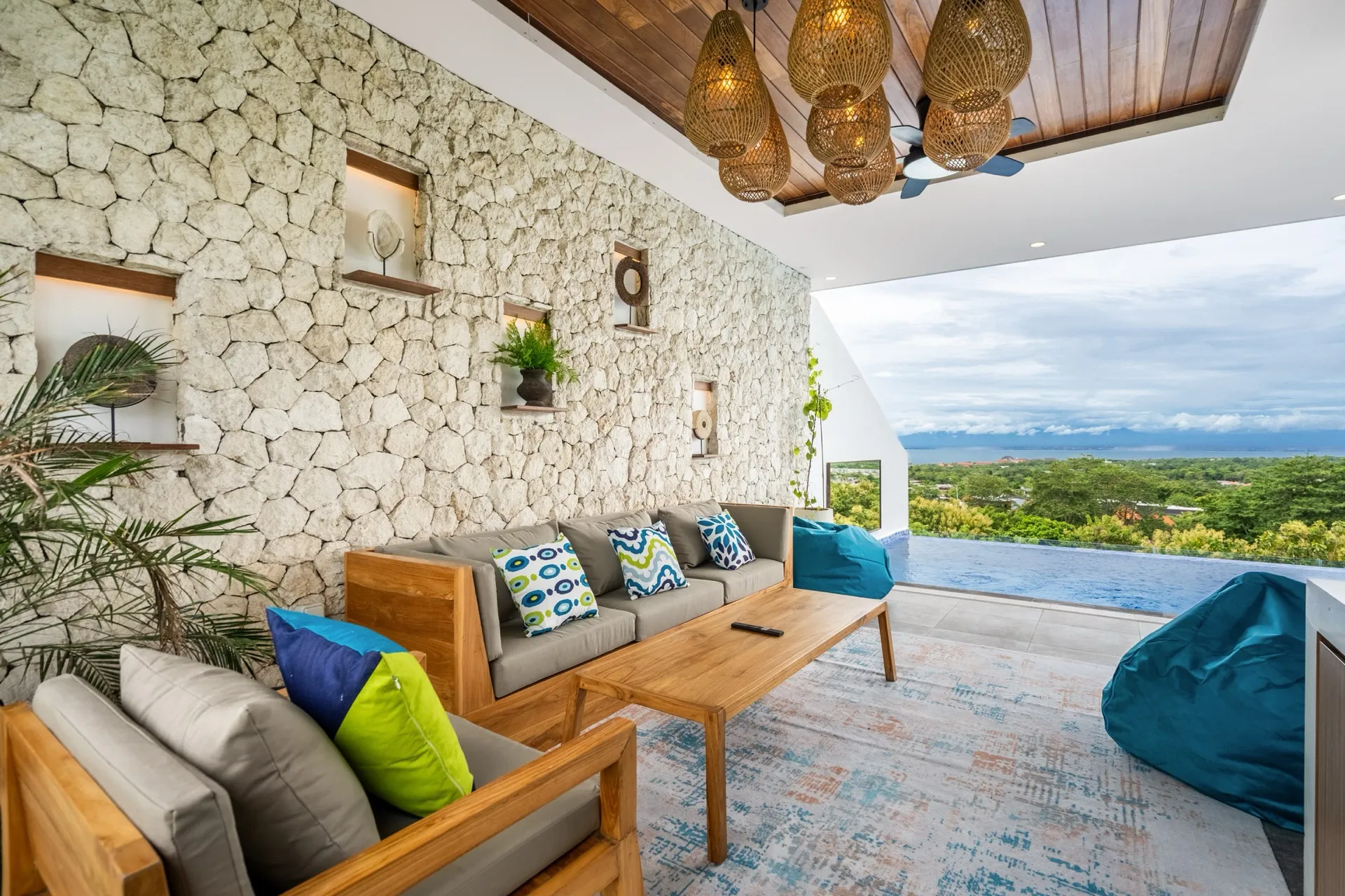 The covered lounge terrace at Villa Pepper, teak sofas against a limestone wall with the infinity pool and coastline beyond